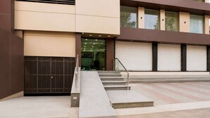 A building with a modern design, featuring a set of stairs leading to a glass entrance, a metal gate, and a series of closed roller shutters. - Classic Simran Raghuveer Para (Rajkot)