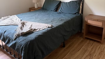 Um quarto com cama, mesa de cabeceira, abajur, piso de madeira e parede com quadros.