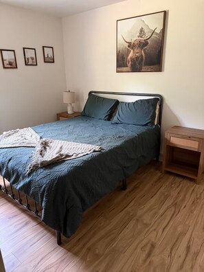 Een slaapkamer met een bed, een nachtkastje, een lamp, een houten vloer en een muur met ingelijste foto's.