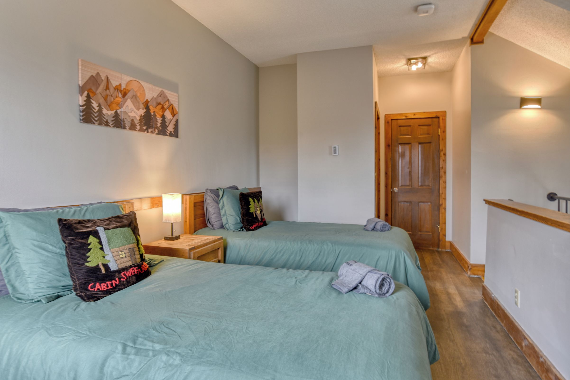 Une chambre avec deux lits, une table de chevet avec une lampe, un tableau au mur et une porte en bois.