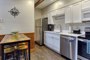 A kitchen with white cabinets, stainless steel appliances, a wooden table with stools, and a wall-mounted compass decoration. - Sunset facing slopeside condo and pet friendly! (Snowshoe)