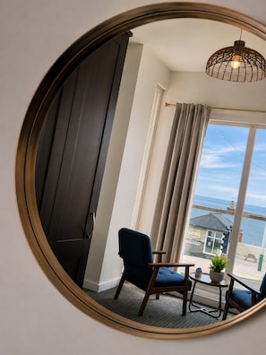 A room with a large mirror, a chair, a small table, and a view of the sea through a window.