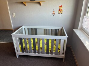 Ein weißes Babybett mit gelber Matratze und Tierprints, vor einer grauen Wand mit einem Fenster zur Rechten.