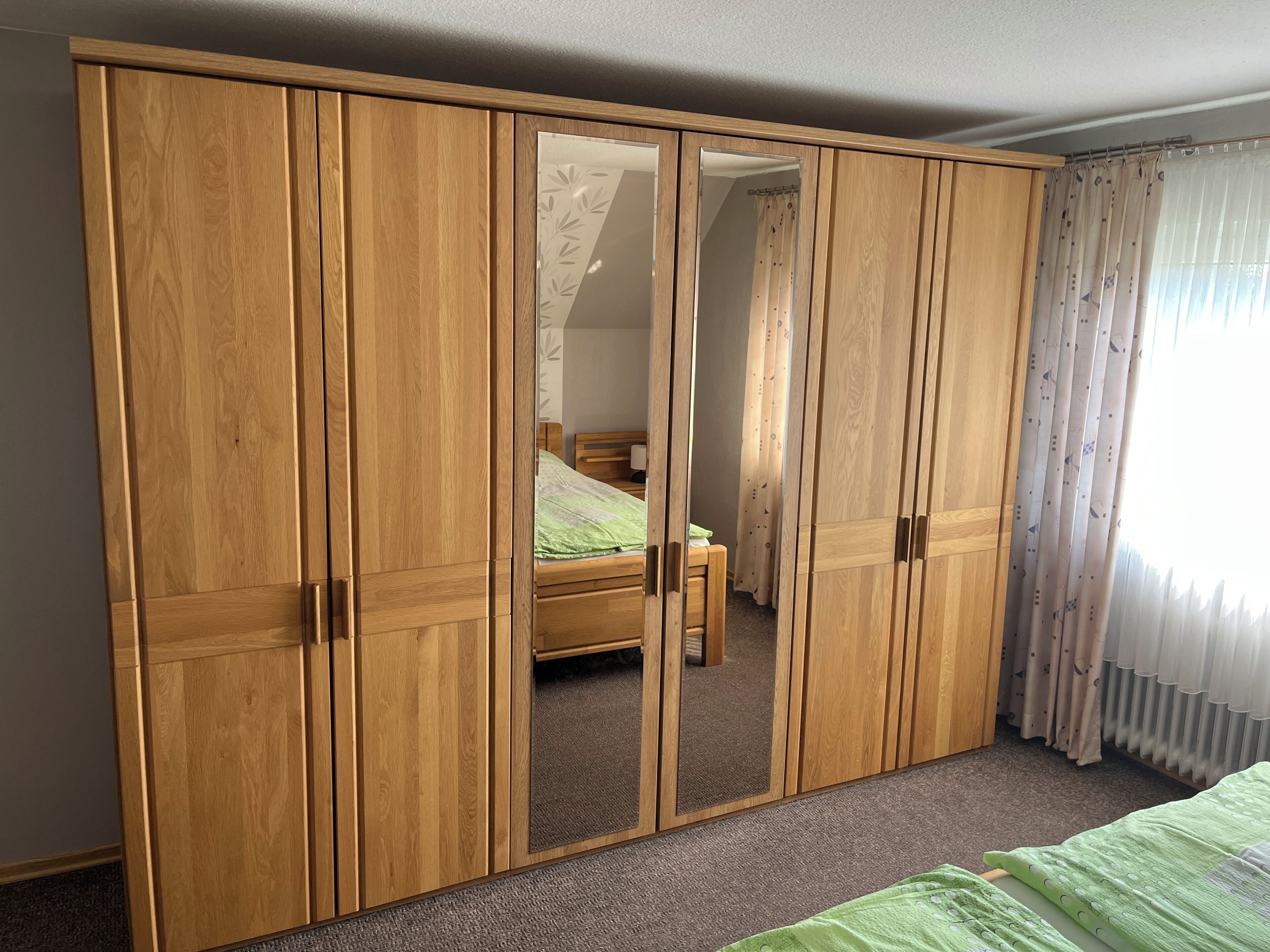 A room with a large wooden wardrobe, a bed with green bedding, and a window with sheer curtains.