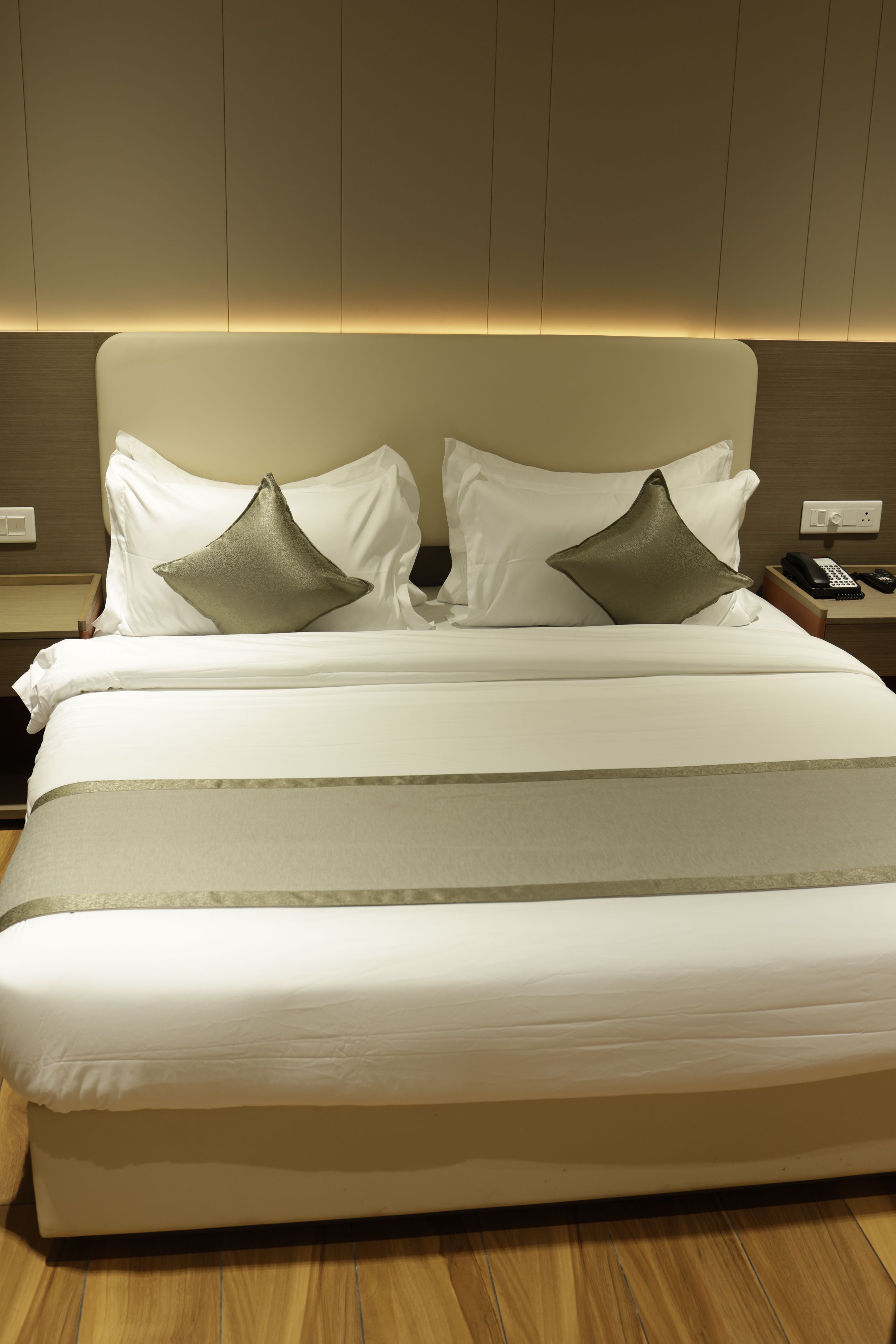 A neatly made bed with white linens and two decorative pillows, a bedside table with a phone and remote control, and a wall-mounted light fixture.