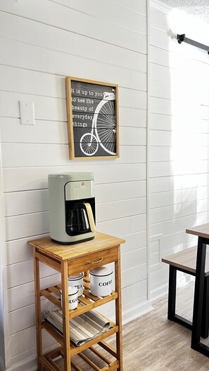 Ein Küchenbereich mit Kaffeemaschine auf einem Holztisch, einem Regal mit beschrifteten Behältern und einem gerahmten Motivationsspruch an der Wand.