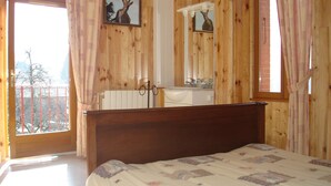 Ein Schlafzimmer mit Holz-Kopfteil, einem Bett mit gemustertem Überwurf, einem Badezimmer mit Spiegel und Waschbecken sowie ein Balkon mit Aussicht.