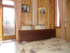 Quarto com cabeceira de madeira, cama com colcha estampada, banheiro com espelho e pia, e varanda com vista.
