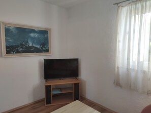 A living room with a flat-screen TV on a wooden stand, a framed painting on the wall, and a window with sheer curtains. - Your Perfect Summer Getaway in Croatia (Krk)
