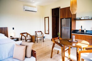 A hotel room with a bed, a sitting area with chairs, a dining table, a kitchen area with a refrigerator, and a hanging light fixture.