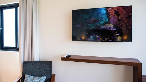 Una habitación con una televisión instalada en la pared, una mesita consola de madera, una silla con cojín y una ventana con cortinas.