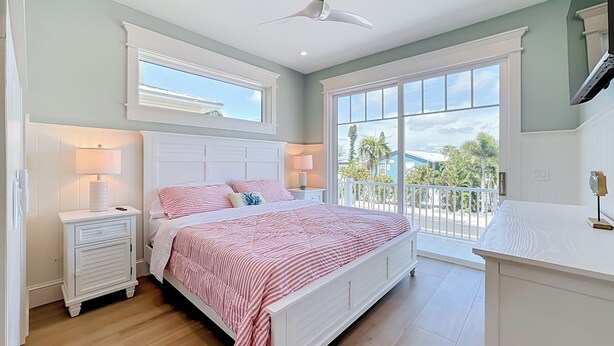 A bedroom with a bed, nightstands, a ceiling fan, and a view of the outdoors. - Coastal Paradise- Brand new 4 bedroom with rooftop elevator (Holmes Beach)