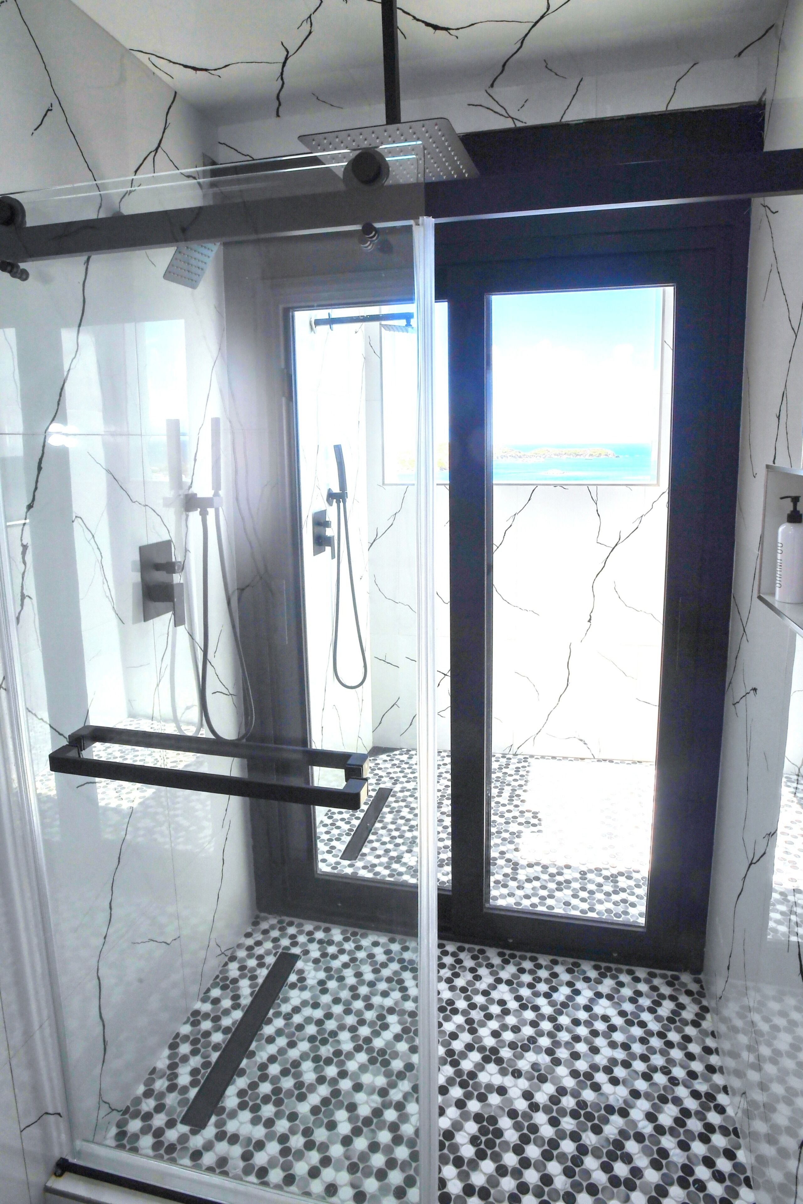 Un espace douche clos par des parois de verre, doté d’un sol carrelé, d’un porte-serviettes et offrant une vue sur la mer par la porte.