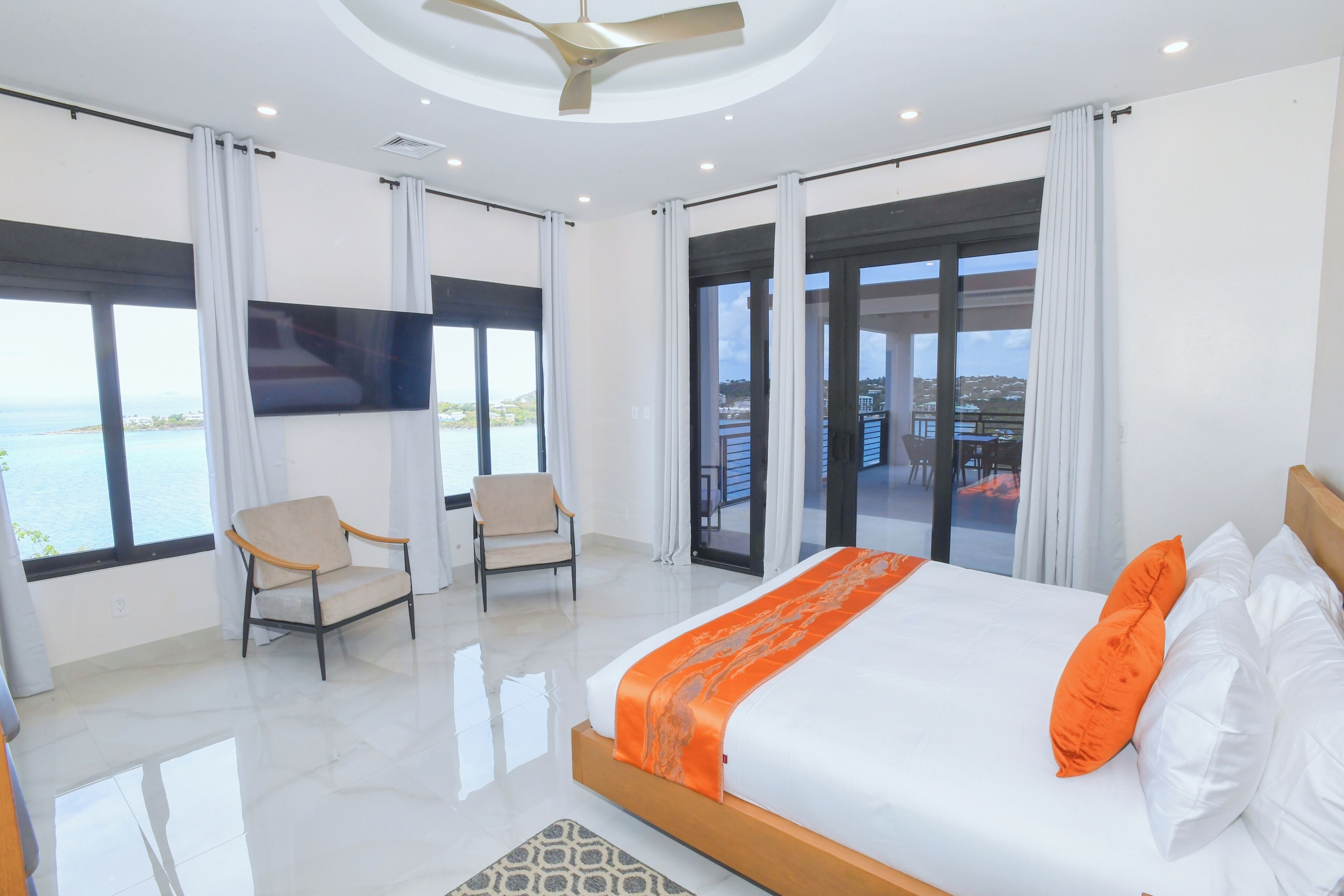 A hotel room with a bed, two armchairs, a TV, and large windows with a view of the sea.