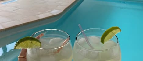 A poolside view with a clear blue pool, two glasses of a drink with lime garnish, and a glimpse of a patio area with outdoor furniture.