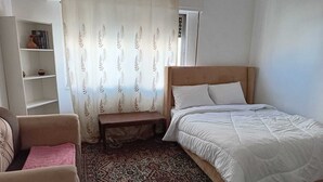 Um quarto com uma cama, um banco, uma cadeira, uma prateleira e um tapete estampado.