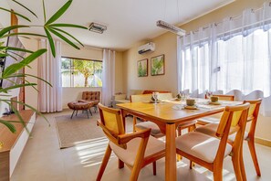 A dining area with a wooden table and chairs, a sofa, and a large window with curtains. - House in Guriri, Cabo Frio – Space & Convenience (Cabo Frio)