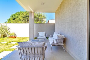 Une terrasse avec deux chaises en métal blanc et une petite table, un mur blanc et une vue dégagée sur un jardin verdoyant.