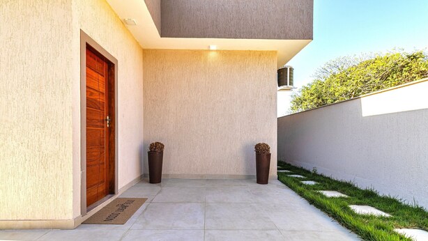 Une entrée moderne avec une porte en bois, deux plantes en pot, un paillasson et un chemin en béton menant à une zone herbeuse.
