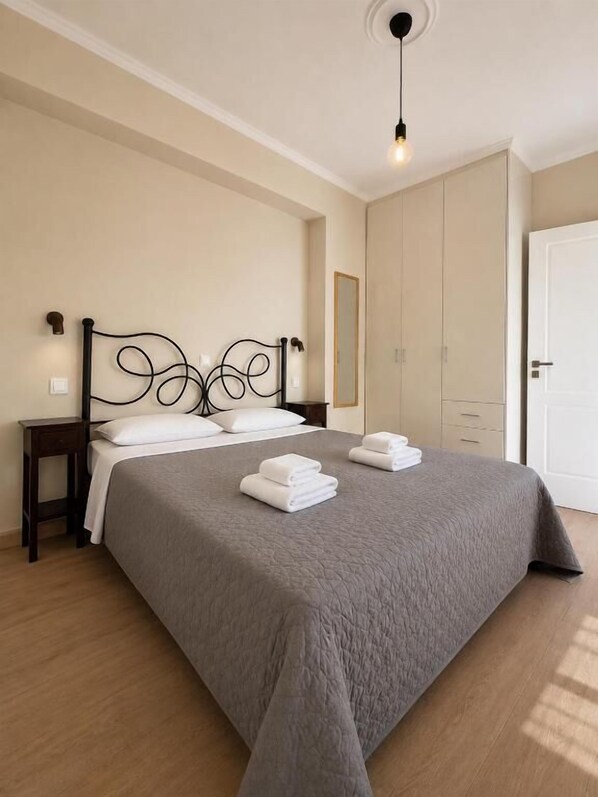 A neatly made bed with a grey bedspread and white pillows, a bedside table with a lamp, and a wardrobe in a room with light-colored walls and wooden flooring. - Angelland Luxury Suites (Nafpaktos)
