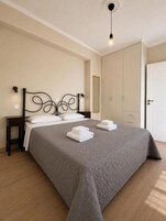 Una cama bien hecha con colcha gris y almohadas blancas, una mesita de noche con lámpara y un armario en una habitación con paredes de tonos claros y suelo de madera.