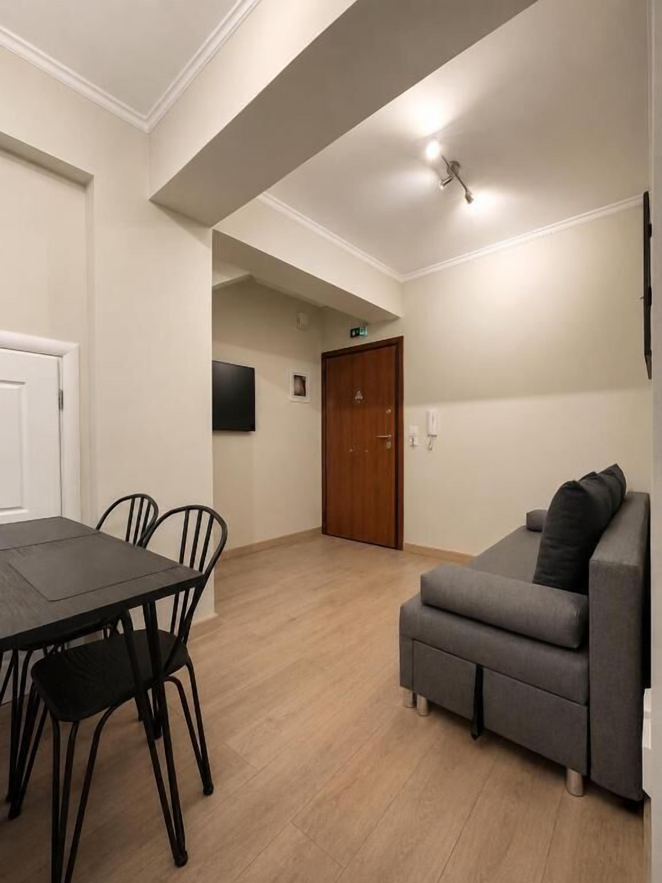 A room with a dining table and chairs, a sofa, a wooden door, and a light fixture on the ceiling.