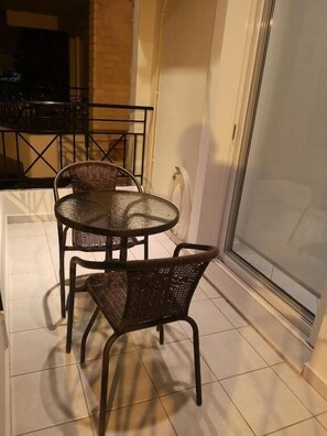 Un balcón con una mesa redonda y dos sillas de mimbre, una puerta de vidrio y piso de baldosas.