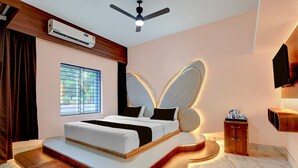 Une chambre à coucher avec un lit doté d’une tête de lit pourvue de lampes en forme d’ailes, un ventilateur de plafond, un appareil de climatisation, une fenêtre avec des stores et une table de chevet avec une bouilloire et des tasses.