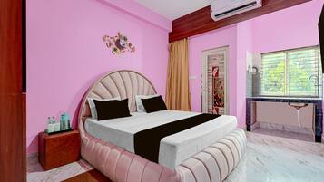 A bedroom with a bed, a bedside table, an air conditioning unit, a window with blinds, and a decorative mirror on the wall.