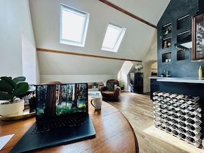 Ein modernes Loft mit schrägem Dach, Dachfenstern, einem Laptop auf einem Holztisch, einem Ledersessel und einem Weinkühlschrank.