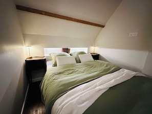 Ein Schlafzimmer mit schrägen Wänden, zwei Nachttische mit Lampen, ein Bett mit grün-weiß gestreifter Bettwäsche und ein Fenster mit Vorhängen.