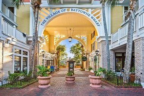 A covered walkway with a sign reading 'The Village of Baytowne Wharf' leading to a building with a brick facade and a balcony. - Luxury Baytowne Wharf- Bahia Breeze -Pool View (Miramar Beach)