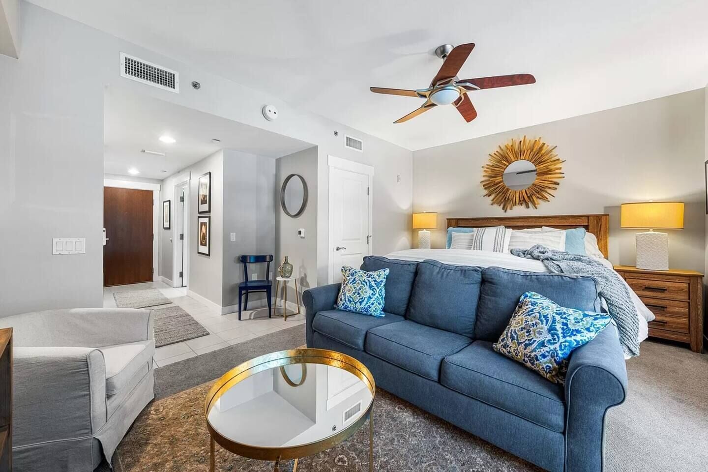 A modern hotel room with a bed, a blue sofa, a round glass coffee table, and a ceiling fan.