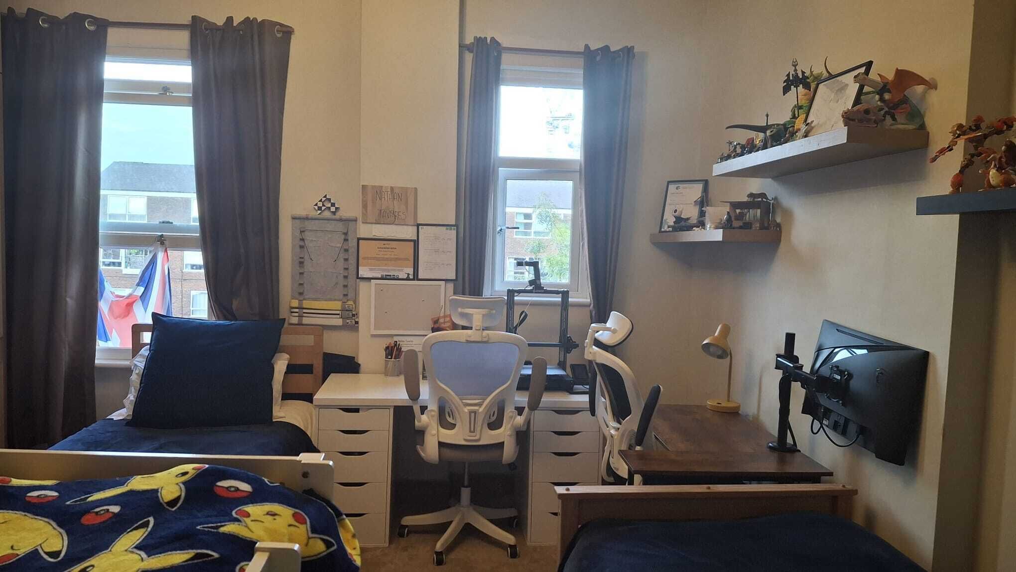 A bedroom with two beds, a desk, a chair, a TV, and a window with curtains.