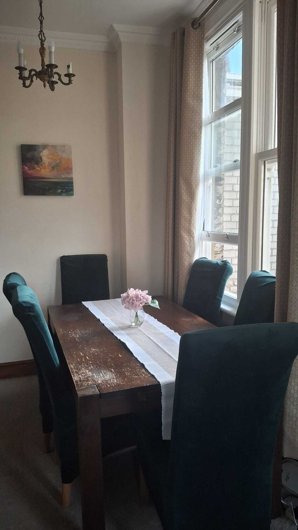 A dining area with a table set for four, chairs, a chandelier, a painting, and a window with curtains.
