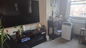 Ein Wohnzimmer mit Fernseher, Bücherregal, kleinem Tisch mit Drucker und Fenster mit Blick auf Gebäude.