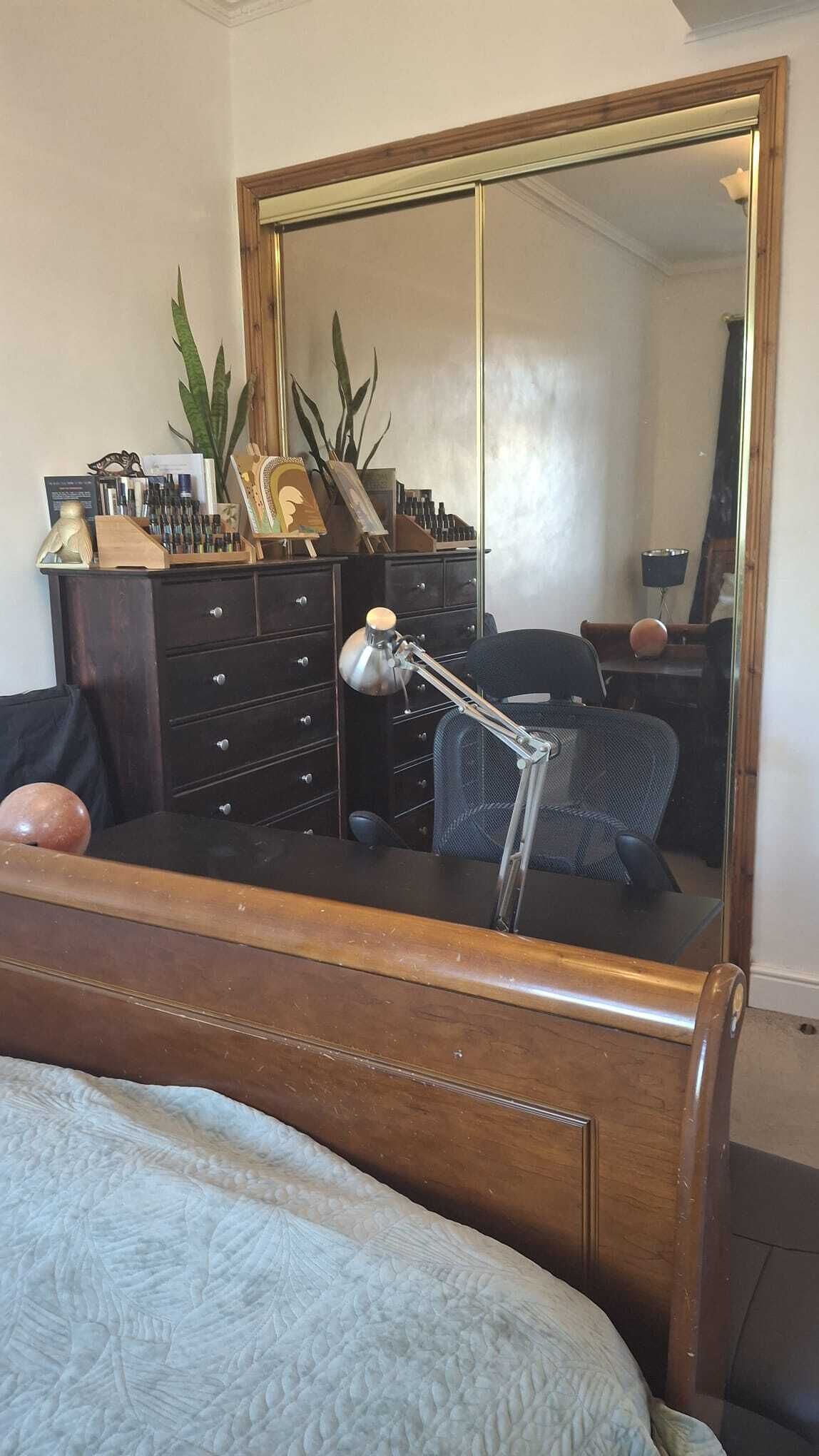 A bedroom with a wooden bed, a dresser with drawers, a mirror, a desk with a chair, and a potted plant.