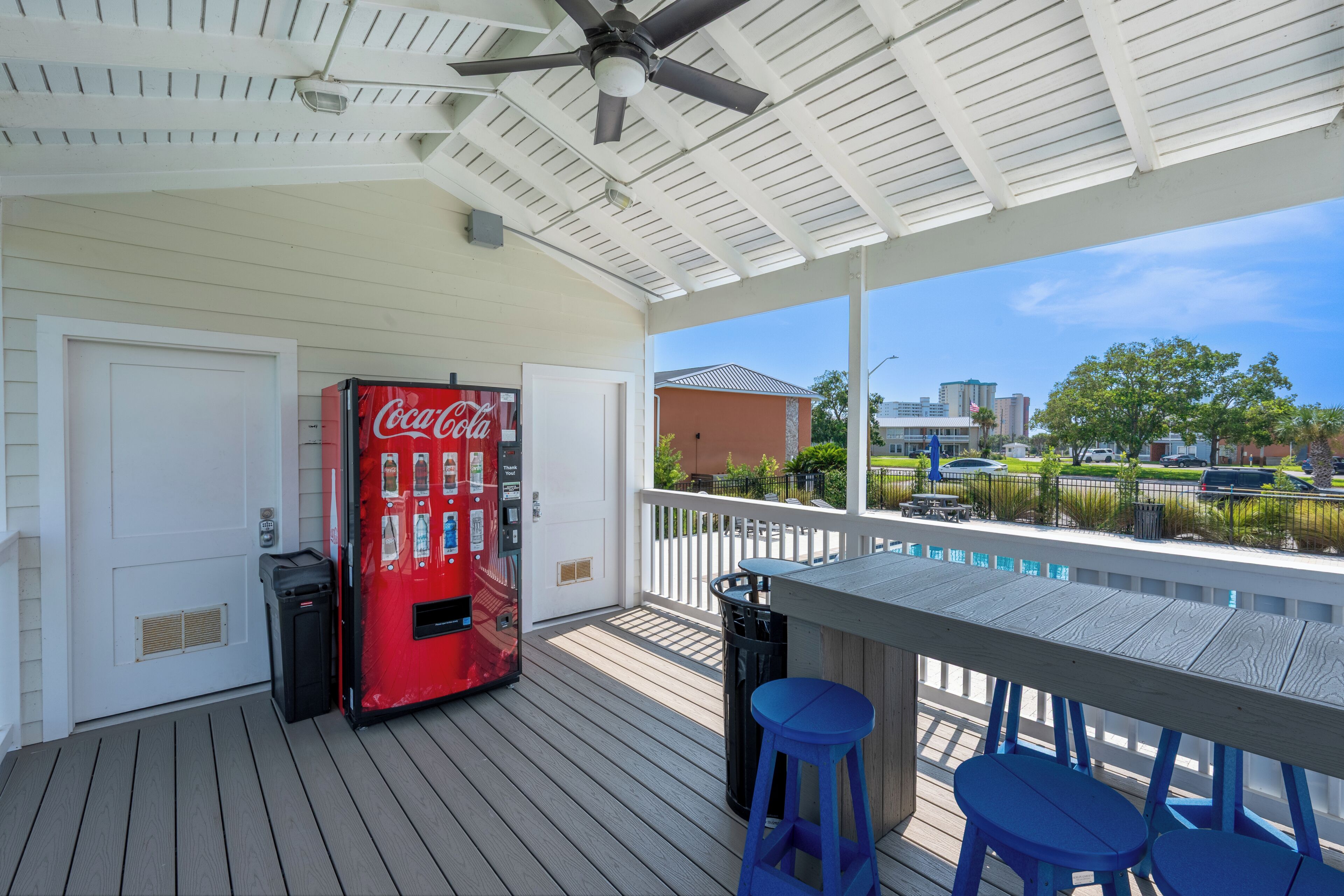 A covered outdoor area with a Coca-Cola vending machine, a table with blue chairs, and a view of buildings and trees.