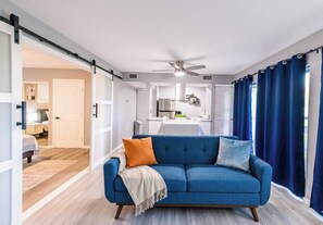 Ein modernes Wohnzimmer mit einem blauen Sofa, einem Deckenventilator und Blick in einen Küchenbereich.