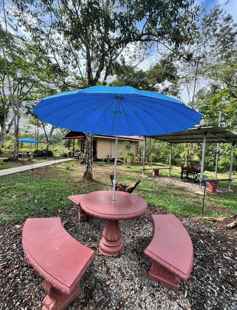 Un jardín con una mesa redonda y bancos, una sombrilla azul y un área techada.