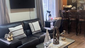 A modern living room with a black leather sofa, a glass coffee table with decorative items, a flat-screen TV mounted on the wall, and a dining area with wooden chairs and a table.