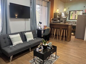 A modern living room with a sofa, a coffee table, and a TV. There is a kitchen area with a bar and stools.