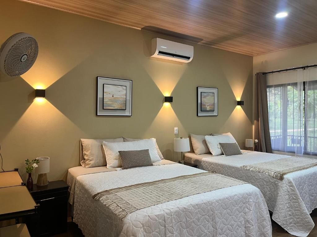 A hotel room with two beds, a ceiling fan, wall-mounted air conditioning, and framed artwork.