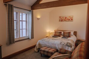 A bedroom with a bed, a nightstand, a wall-mounted lamp, a window with blinds, and a painting on the wall.