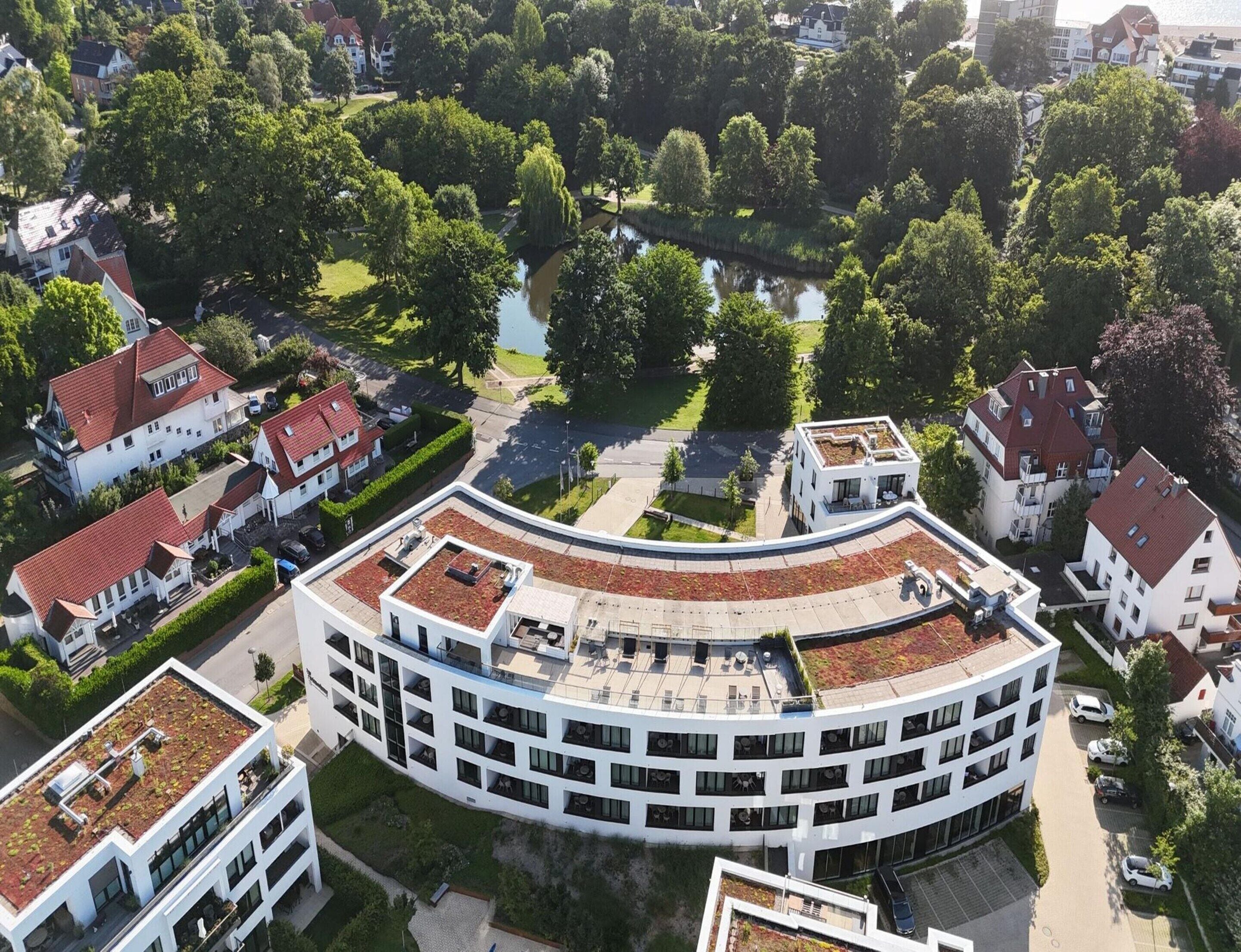 Um edifício moderno com terraço curvo no telhado, cercado por vegetação exuberante e um pequeno lago.