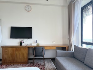 A hotel room with a bed, a sofa, a desk with a flat-screen TV, a chair, a clock on the wall, and a window with curtains.