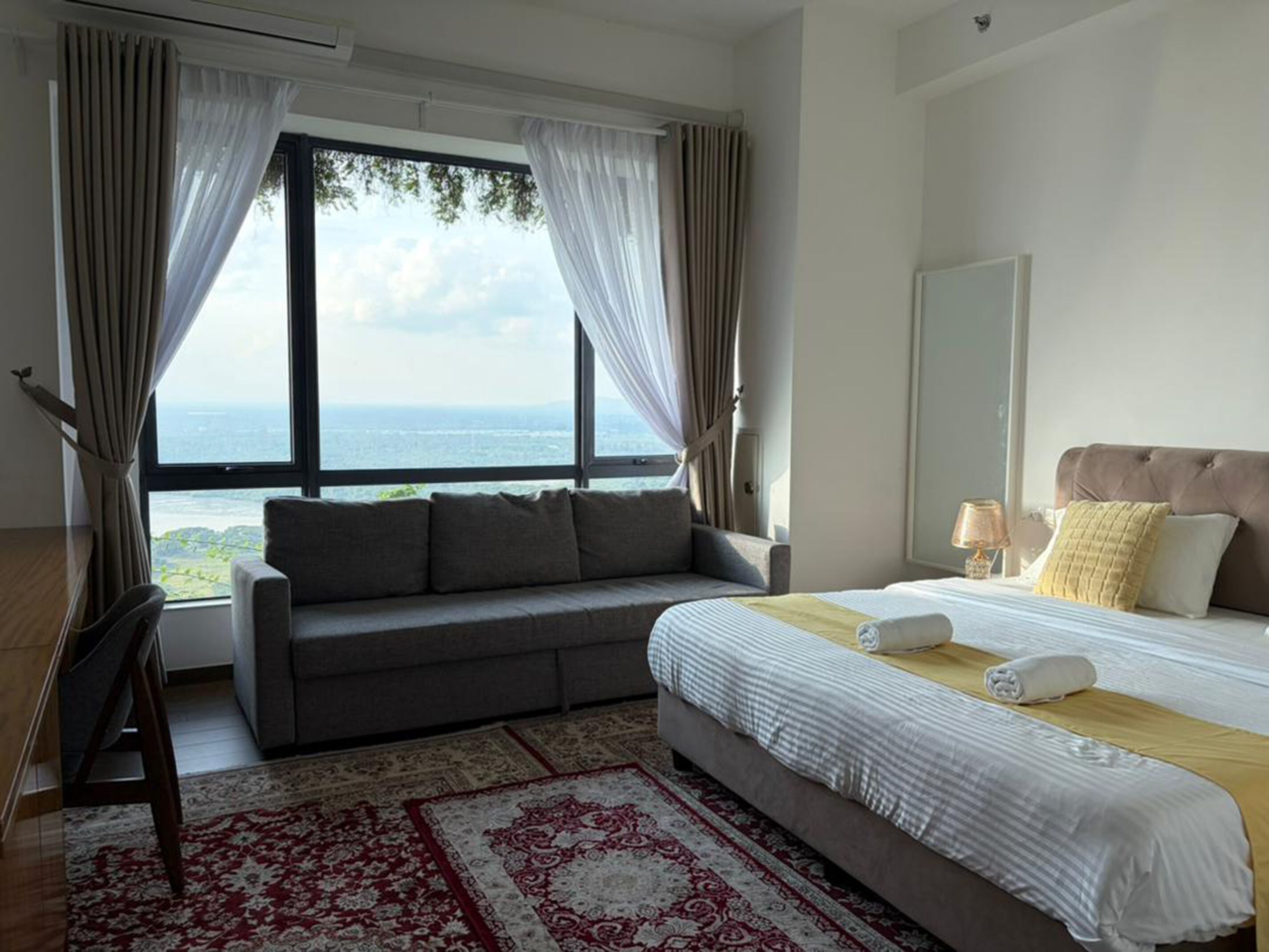 A hotel room with a large window, a sofa, a bed with a headboard, and a patterned rug.