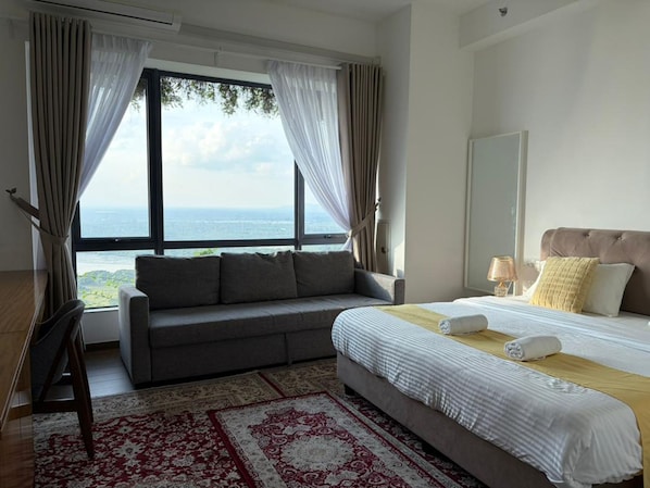 A hotel room with a large window, a sofa, a bed with a headboard, and a patterned rug.
