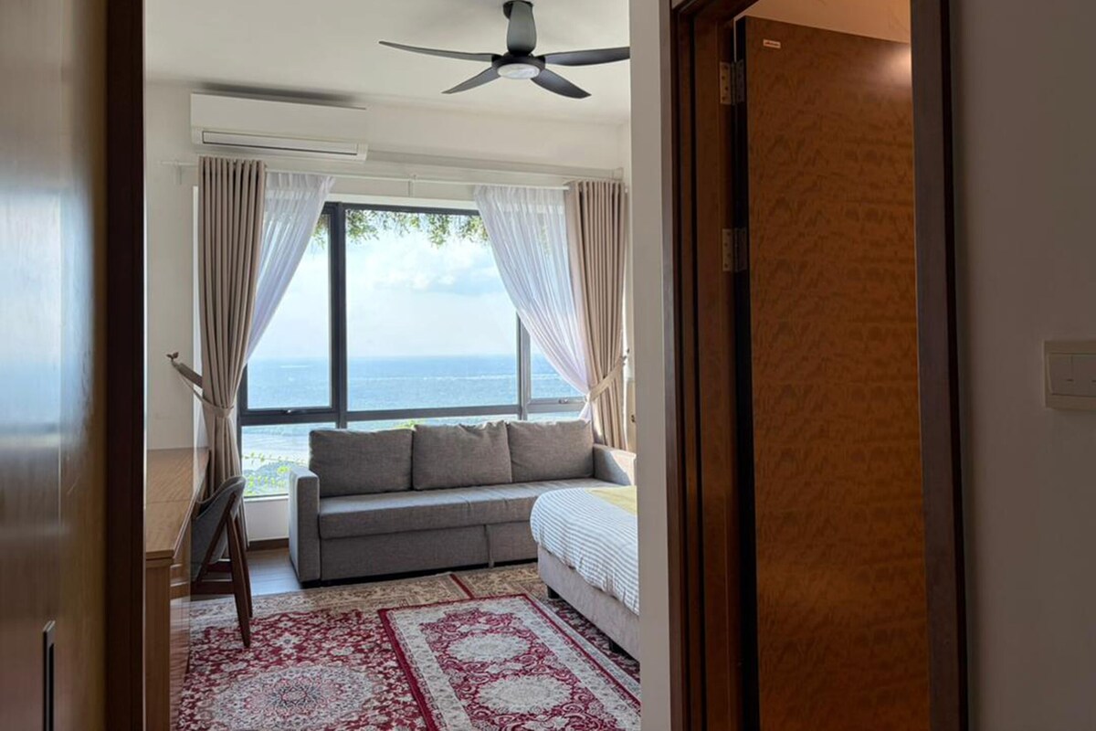 A room with a sofa, a ceiling fan, and a view of the sea through large windows.