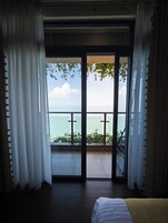 A room with a bed, a view of the sea through a glass door, and a balcony with plants.